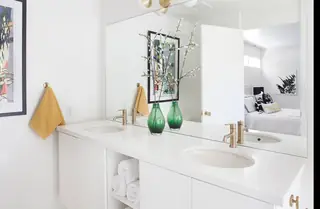 Pebble Beach Drive, Palm Springs: A bright, modern bathroom features a white double vanity with gold faucets, green glass vases, and a large mirror reflecting an adjacent bedroom