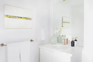 Pebble Beach Drive, Palm Springs: A bright minimalist white bathroom vanity features a sleek gold faucet, light celadon vases with orchids, and framed abstract art on the wall