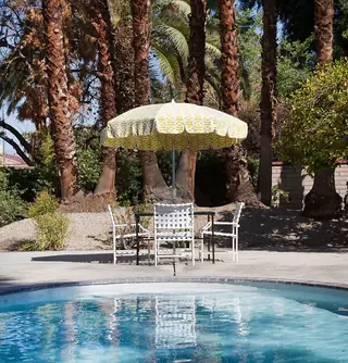 Pebble Beach Drive, Palm Springs: A poolside patio features a patterned yellow fringe umbrella and a white woven dining set set against a backdrop of palm trees