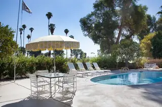 Pebble Beach Drive, Palm Springs: A sunny poolside patio featuring a yellow patterned umbrella over an outdoor dining table, lined with white loungers under palm trees