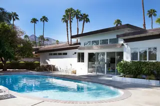 Pebble Beach Drive, Palm Springs: A mid-century modern white house exterior featuring a backyard swimming pool, concrete patio, and tall palm trees against a mountain backdrop