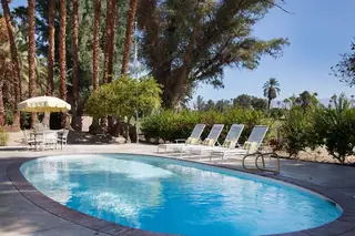Pebble Beach Drive, Palm Springs: A sunlit outdoor swimming pool area features white lounge chairs with yellow accent pillows, a shaded dining set