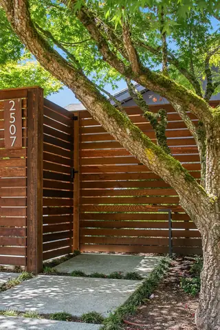 A modern wooden slat fence with a vertically mounted house number plaque stands along a concrete paver walkway beneath a moss-covered tree.