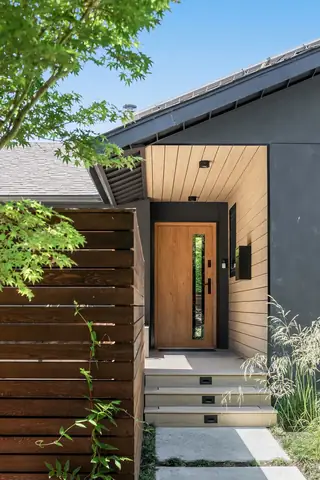 A modern home entrance features a light wood-lined alcove porch, a matching wooden front door, and dark gray exterior siding framed by a dark wood privacy fence.