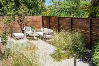 A modern outdoor patio surrounded by a dark wood slatted privacy fence features contemporary lounge seating set among lush ornamental grasses.