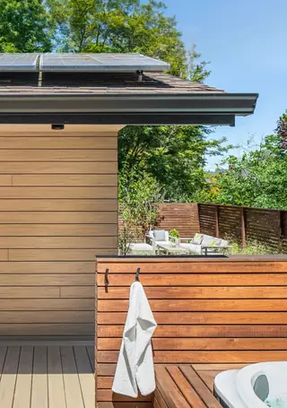 A modern outdoor deck features horizontal wood siding, a hot tub area with a hanging white towel, and patio furniture set in the background garden.
