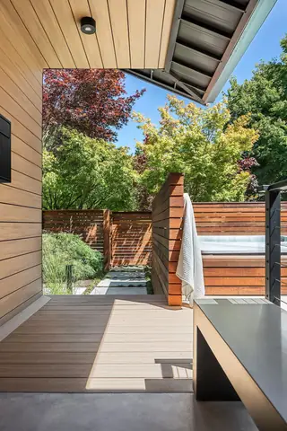 A modern covered deck offers a view of a private backyard patio with a hot tub, warm wooden privacy screens, and lush trees.