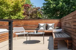 A modern outdoor deck featuring built-in wooden bench seating, a round black coffee table, and a white patterned accent chair against a horizontal wood slat fence.