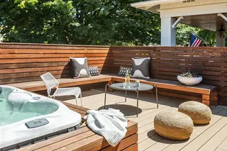 A modern outdoor deck features built-in wooden bench seating around a small coffee table, a hot tub, and woven poufs.