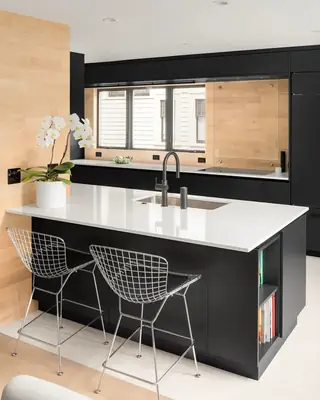 A modern kitchen features a sleek black island topped with white quartz, paired with chrome wire barstools and warm wood wall paneling.