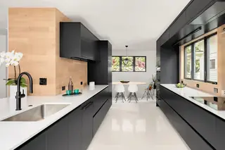 A sleek modern kitchen features matte black cabinetry, light wood wall paneling, white quartz countertops, and a dining area in the background.