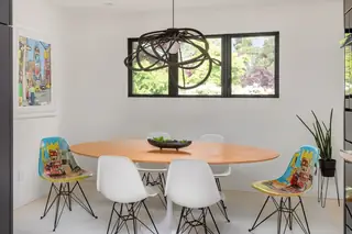 A modern dining area features an oval wooden table surrounded by white and graphic art-printed Eames-style chairs beneath a sculptural dark wire pendant light.