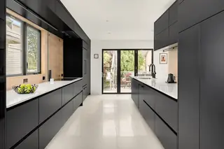 A sleek modern galley kitchen features matte black cabinetry, white countertops, light tile flooring, and glass patio doors leading outside.