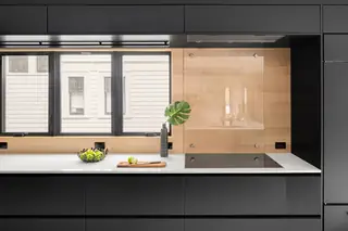 A sleek modern kitchen features matte black cabinetry, a white countertop with an induction cooktop, and warm wood accents around the backsplash and windows.