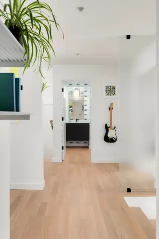 A bright modern hallway with light hardwood floors features an electric guitar mounted on a white wall leading into a bathroom with graphic wallpaper.