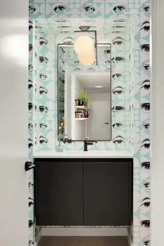A modern powder room features whimsical light blue graphic eye wallpaper surrounding a black vanity cabinet and a thin-framed mirror.