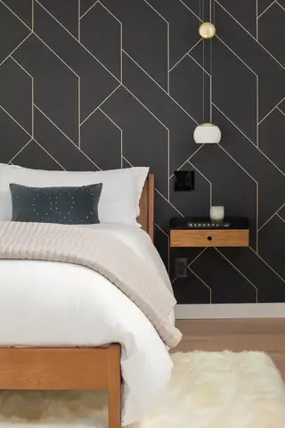 A modern bedroom features a wooden bed frame dressed in white bedding set against a dark charcoal wallpaper with geometric gold lines.