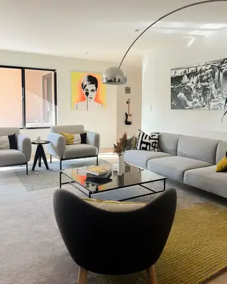 A bright modern living room features a grey sectional sofa, two light grey armchairs, a black coffee table, and vibrant pop art on the wall.