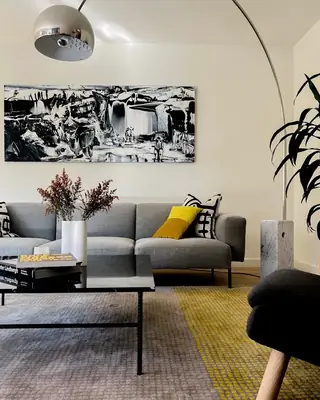 A modern living room features a light gray sectional sofa beneath a dramatic black-and-white abstract painting, complemented by an arc floor lamp and a low black coffee table.