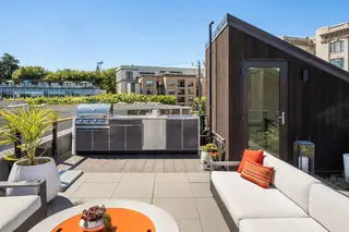 A modern rooftop deck features a stainless steel outdoor kitchen station, stylish sofa with orange accent pillows, and urban skyline views.