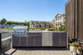 An outdoor rooftop kitchen features sleek grey cabinetry, an integrated stainless steel barbecue grill, and potted flowers on a wooden deck.
