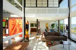 The Aqua Lair, Lake Sammamish: A sunlit mid-century modern open-plan living room features high ceilings, floor-to-ceiling windows, a brown sofa, and a bold orange front entrance