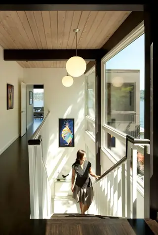 The Aqua Lair, Lake Sammamish: A woman walks down a dark wood staircase in a modern home featuring wood-paneled ceilings, globe pendant lights, and floor-to-ceiling windows