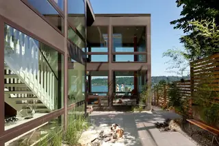 The Aqua Lair, Lake Sammamish: A modern lakeside residential building with floor-to-ceiling glass windows and a stone patio courtyard where a dog rests near a wooden bench