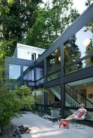The Aqua Lair, Lake Sammamish: A woman relaxes on a modern paved courtyard patio outside a two-story glass-fronted mid-century home