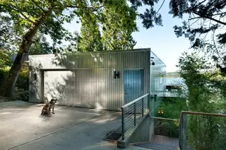 The Aqua Lair, Lake Sammamish: A modern silver metal-clad garage structure overlooks a lake surrounded by trees, with a dog sitting on the concrete driveway