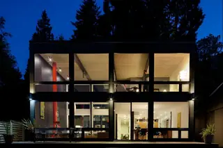 The Aqua Lair, Lake Sammamish: A two-story modern glass house illuminated at night, revealing a stylish wood-paneled ceiling, red structural pipe, and open-plan interior
