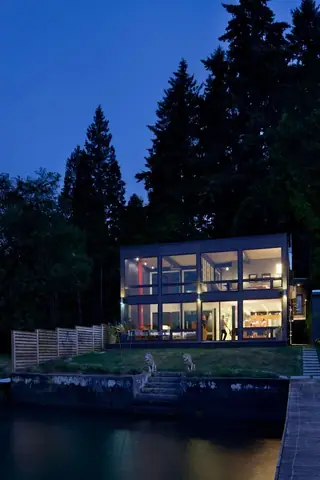 The Aqua Lair, Lake Sammamish: A dusk view of a modern two-story glass house lit from within, set on a waterfront lawn bordered by tall trees