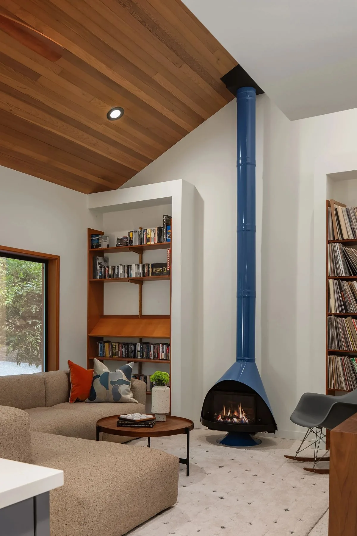 A mid-century modern living room features a striking blue retro fireplace, vaulted wood ceiling, built-in bookshelves, and low beige sectional sofa.