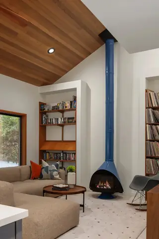 A mid-century modern living room features a striking blue retro fireplace, vaulted wood ceiling, built-in bookshelves, and low beige sectional sofa.