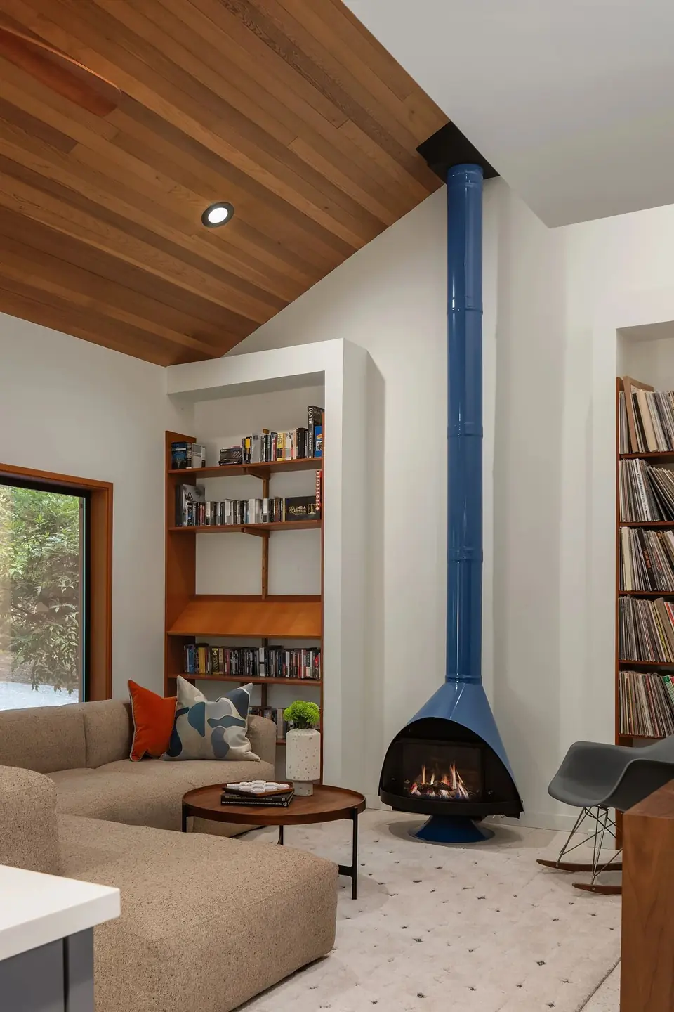 A mid-century modern living room with a striking blue retro fireplace, vaulted wood ceiling, built-in bookshelves, and a low beige sectional sofa.