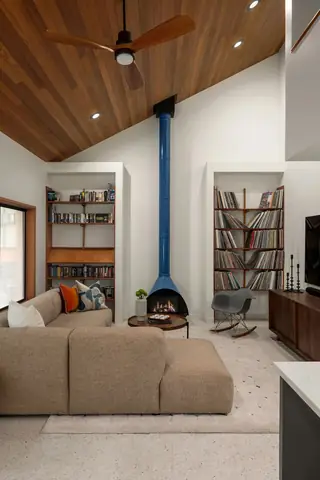A mid-century modern living room features a vaulted wood ceiling, built-in shelving filled with records and books, and a tall blue freestanding fireplace.