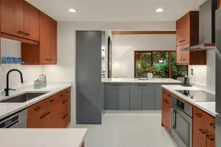 A modern kitchen features rich wood cabinetry combined with sleek grey base cabinets, bright white countertops, and a large window looking out to greenery.