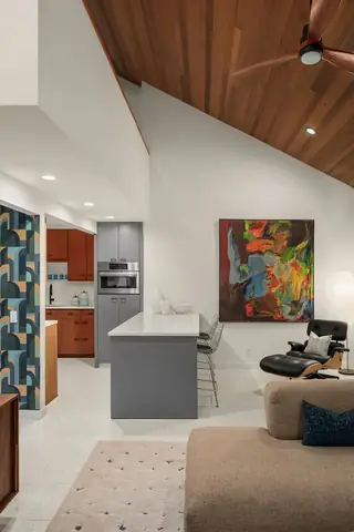 An open-concept mid-century modern home interior featuring a vaulted wood-paneled ceiling, grey kitchen island, and living space with an Eames lounge chair and large abstract artwork.