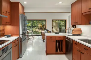 A modern mid-century style kitchen features warm wood cabinetry, white countertops, stainless steel appliances, and a view into a light-filled living area and backyard.