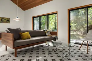 A mid-century modern living room features a wood-framed sofa with a yellow accent pillow, a round glass coffee table, and large sliding glass windows framing a lush forest view.