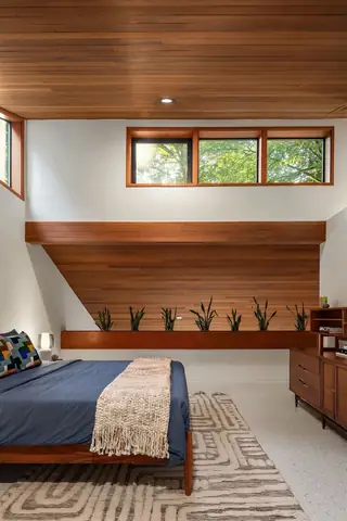 A mid-century modern bedroom featuring a wood-paneled ceiling, a low platform bed with navy bedding, and a row of potted snake plants along a wooden ledge.