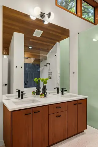 A contemporary bathroom features a warm wood double vanity with white countertops, matte black fixtures, and a large frameless mirror reflecting a sloped wood ceiling.