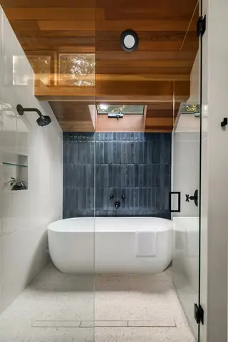 A modern bathroom features a freestanding white bathtub inside a glass shower enclosure, highlighted by a dark blue vertical tile accent wall and a wood-paneled ceiling with a skylight.