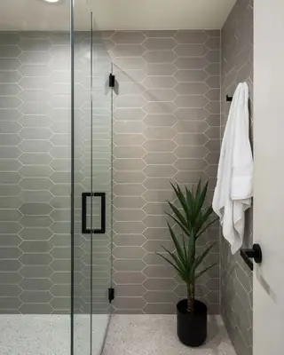 A minimalist bathroom interior showcases grey picket wall tiles, a glass shower enclosure with matte black hardware, terrazzo flooring, and a potted plant.