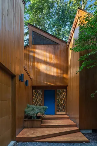 A modern wooden home entrance features vertical timber siding, a vibrant blue front door with decorative glass sidelights, and wooden steps.
