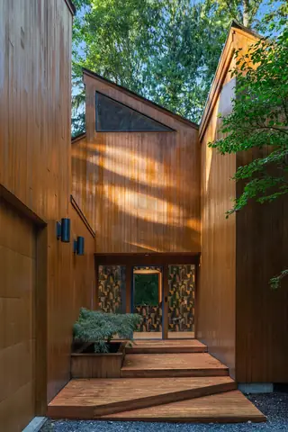A warm wood-clad architectural entryway features asymmetrical wooden steps leading to a patterned glass front door under slanting sunlight.