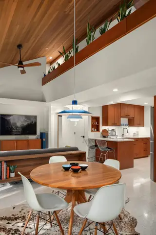 A mid-century modern dining room features a round wood table with white shell chairs beneath a blue PH 5 pendant light, opening to a kitchen background.